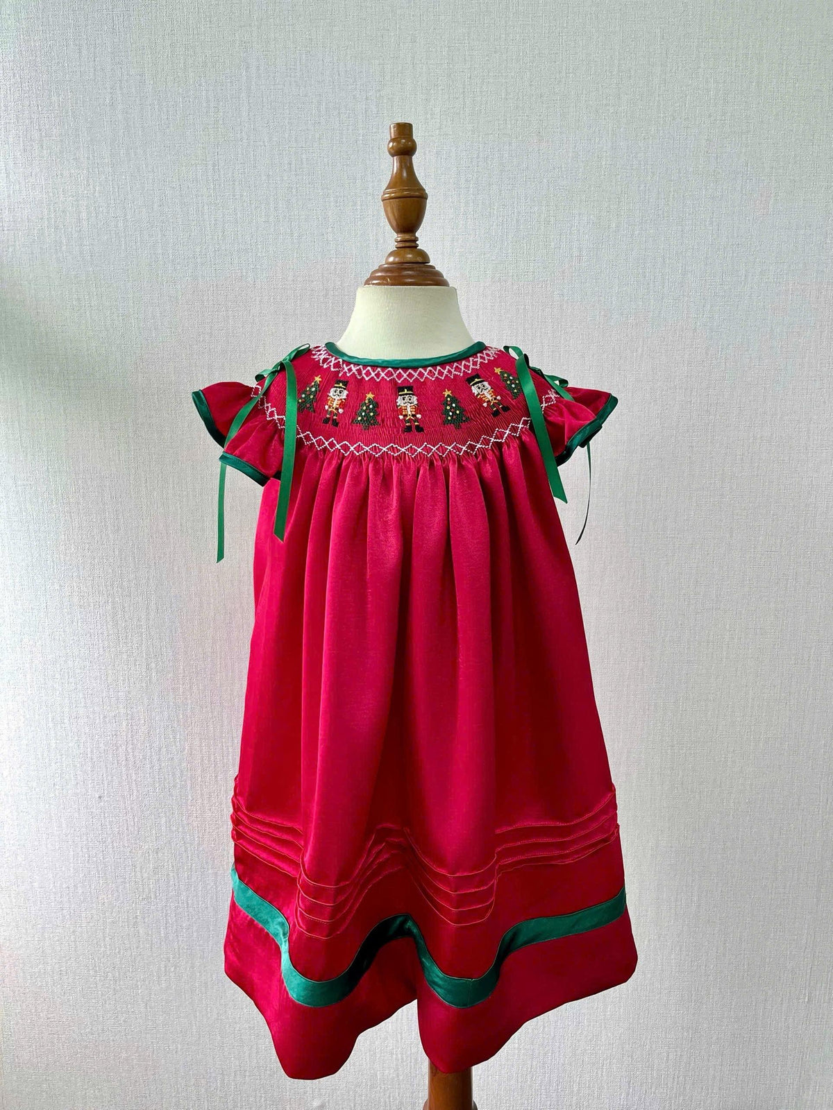 Front view of the red Nutcracker dress on a dress form against a pale wall, with green shoulder bows and hem ribbon.