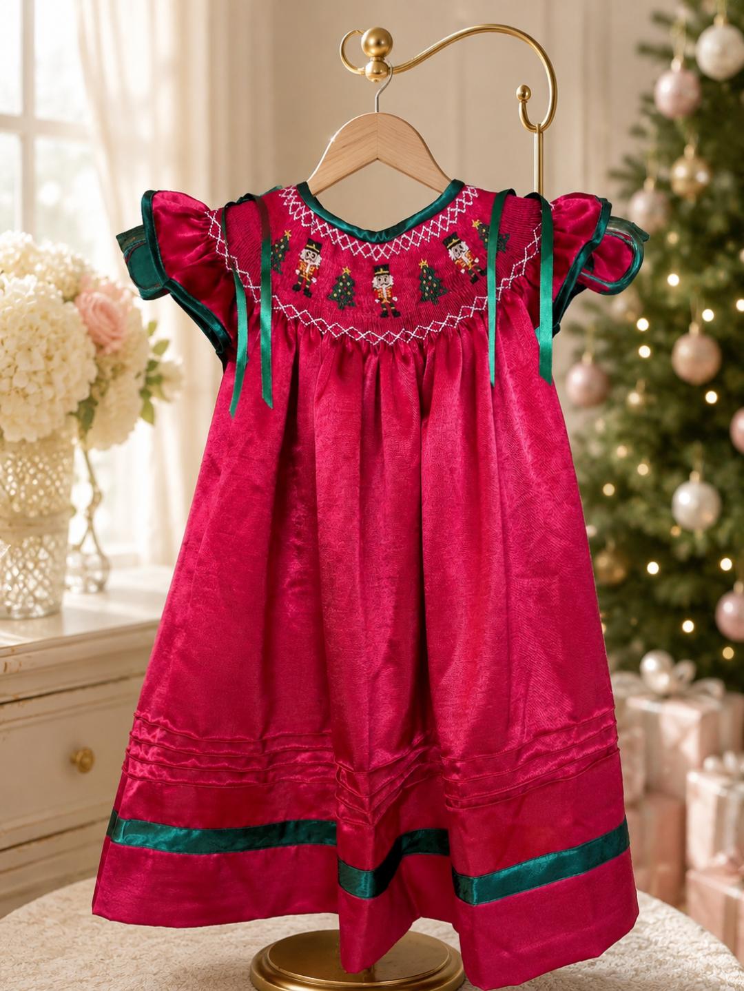 Red satin Nutcracker dress on a hanger beside a Christmas tree, showing embroidered smocking and green ribbon trim.